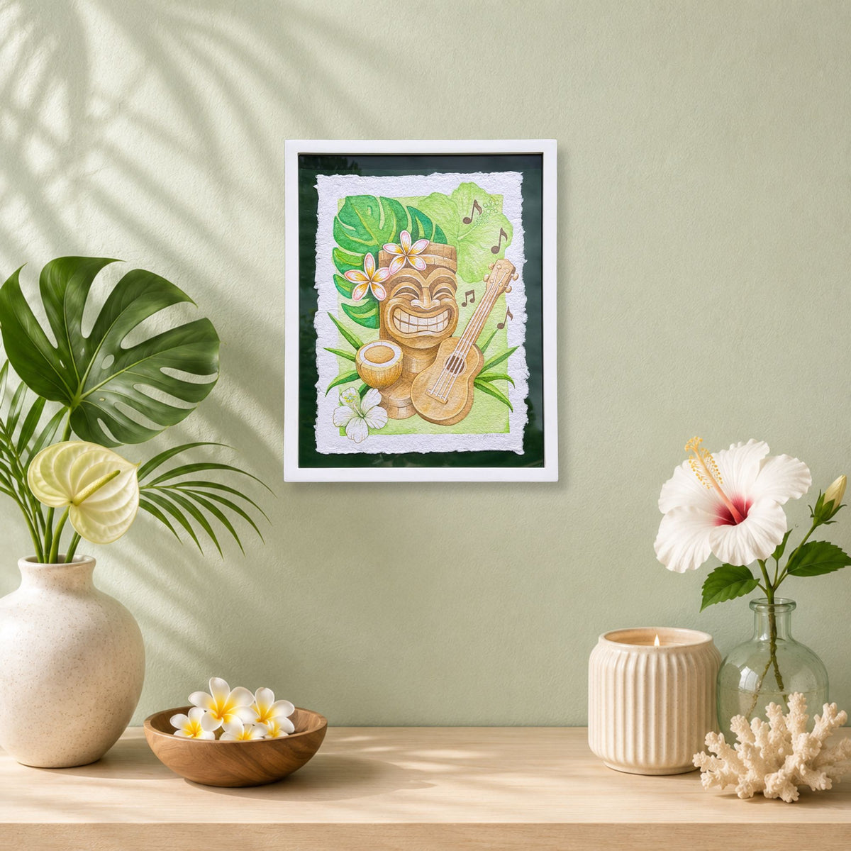 Luxury Hawaiian home with artwork of a tiki with a ukulele on a wall with decorative plants and vases. Framed artwork of a tiki, ukulele, and tropical flowers with leaves on a green background. Aloha Mele, watercolor painting on handmade paper, by Liliane Avalos, Kauai Hawaii 2026.