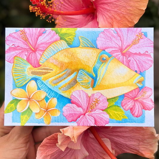 Colorful illustration of a fish surrounded by tropical flowers held against a floral background. Humu Harmony Hawaii art greeting card featuring humuhumunukunukuapua'a reef fish artwork by Liliane Avalos