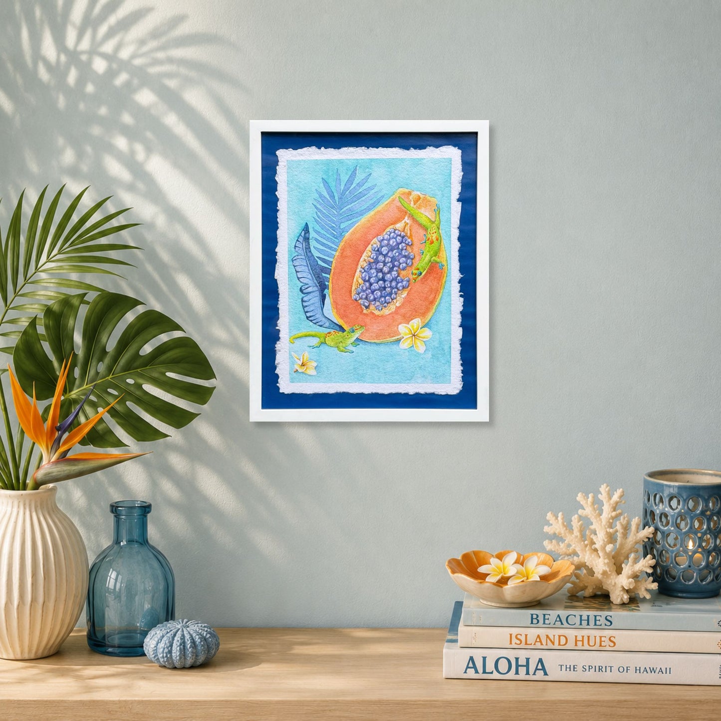 Luxury Hawaiian home with artwork of a Sunrise papaya with leaves on a wall above a table with decorative items. Framed artwork of tropical plumeria flower with palm leaves on a blue background. Good Morning Geckos, watercolor painting on handmade paper, by Liliane Avalos, Kauai Hawaii 2026. 