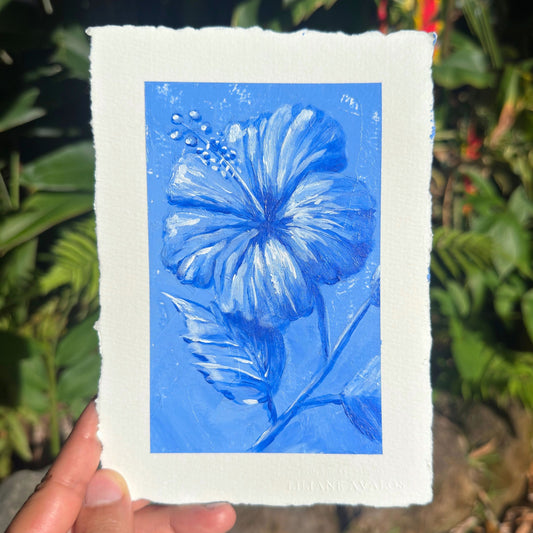 A hand holds a square art print featuring a blue hibiscus flower painted on textured white paper, with a blurred green garden background. Liliane Avalos Kauai Hawaii 2026