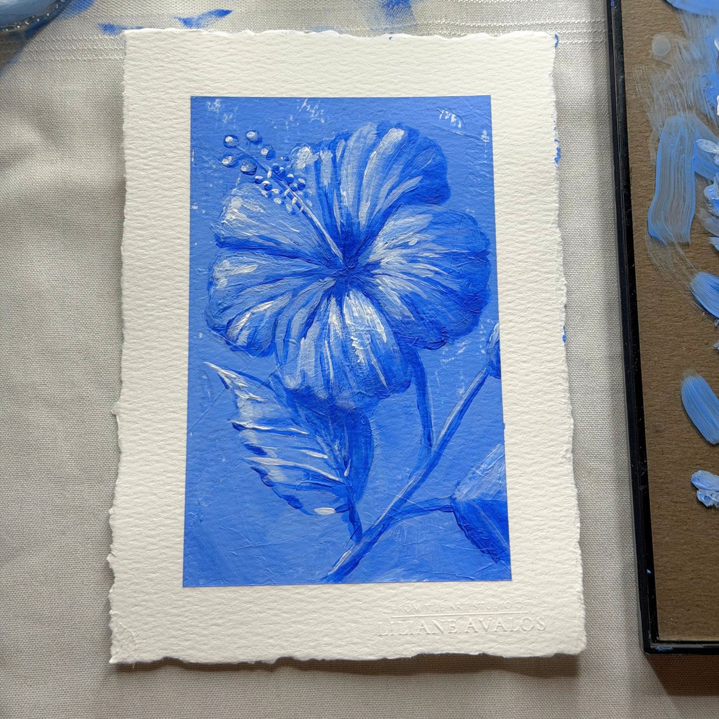A square art print featuring a blue hibiscus flower and leaves on textured white paper with a white border, photographed on a light fabric surface next to a paint palette. Liliane Avalos Kauai Hawaii 2026