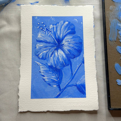 A square art print featuring a blue hibiscus flower and leaves on textured white paper with a white border, photographed on a light fabric surface next to a paint palette. Liliane Avalos Kauai Hawaii 2026