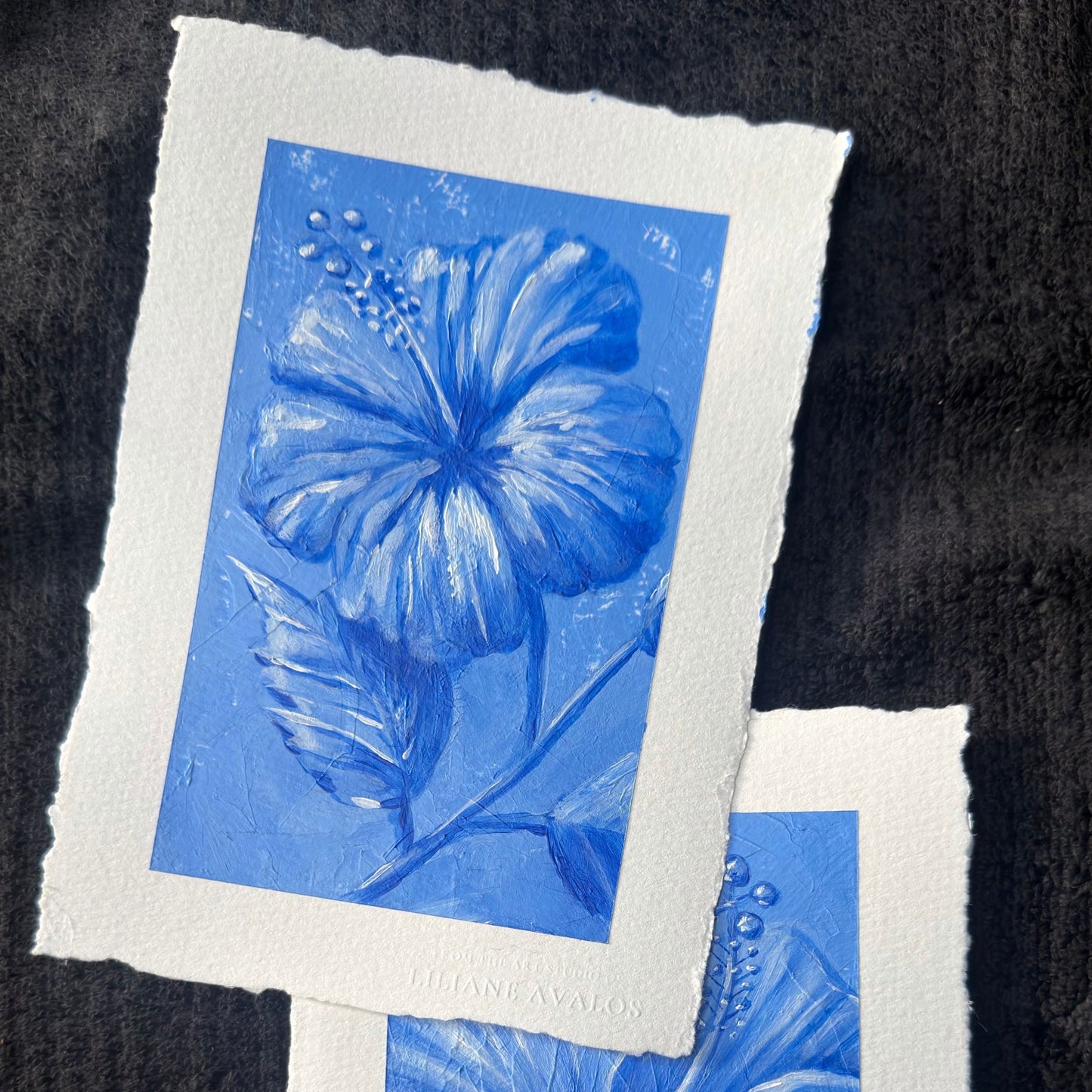 A square art print with a white textured border shows a monochrome blue floral illustration on a dark textured background. A second overlapping print is partially visible at the bottom right. Liliane Avalos Kauai Hawaii 2026