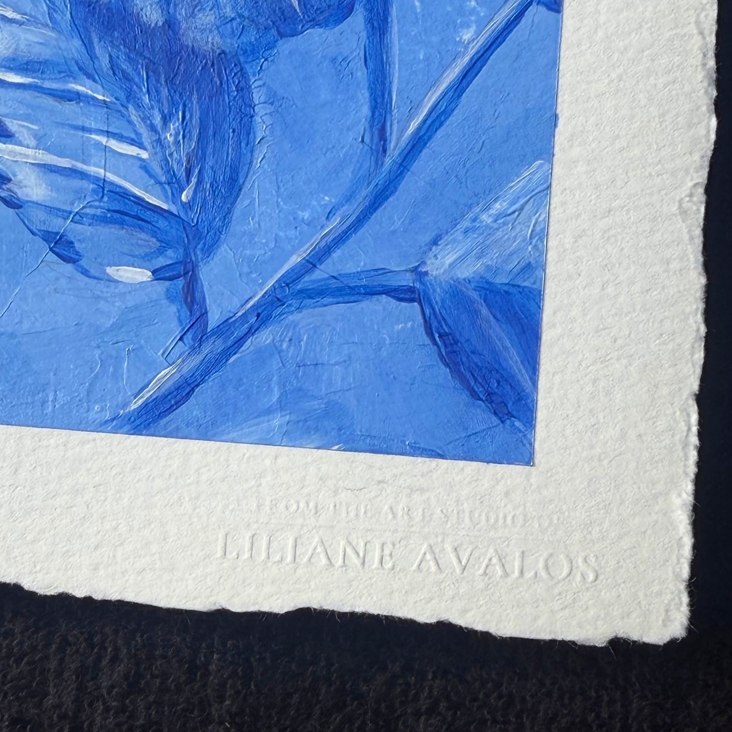 Close-up of a textured white art print with a blue painted floral design and embossed text reading 'LILIANE AVALOS' on the lower margin. Liliane Avalos Kauai Hawaii 2026