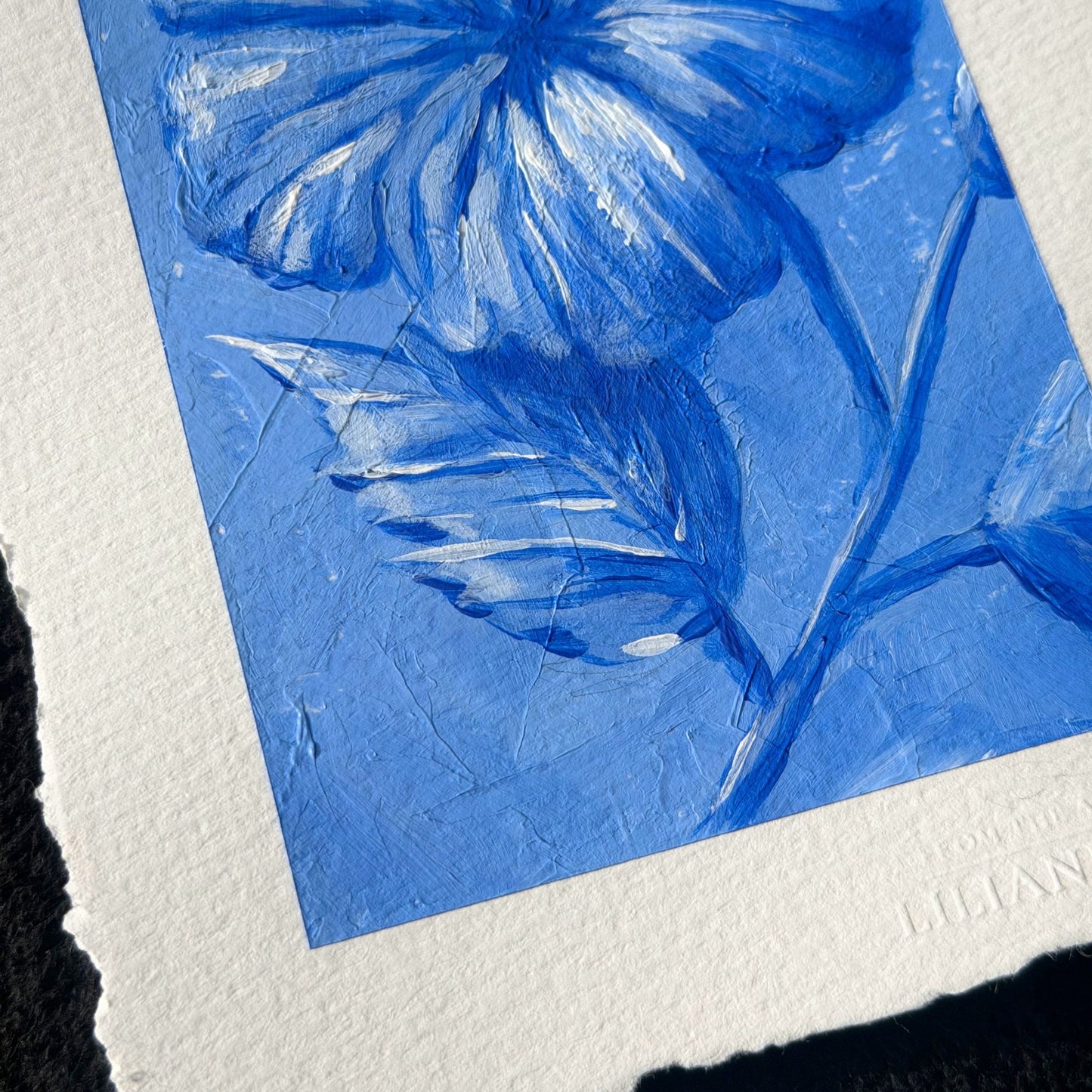 A close-up of a textured blue floral art print on white paper, showing painted flower petals and leaves with visible brush and paper texture Liliane Avalos Kauai Hawaii 2026