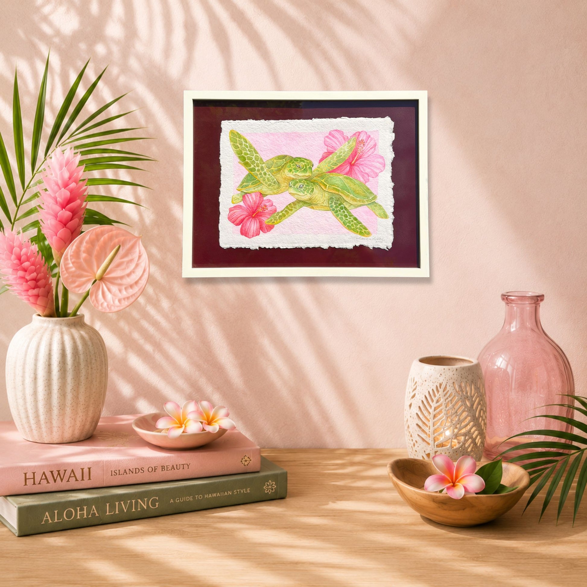 Decorative setup with a framed artwork of a green bird and pink flowers, books, and vases on a wooden surface. watercolor painting on handmade paper of a green sea turtles with pink flowers on a textured background by artist Liliane Avalos Kauai Hawaii 2026