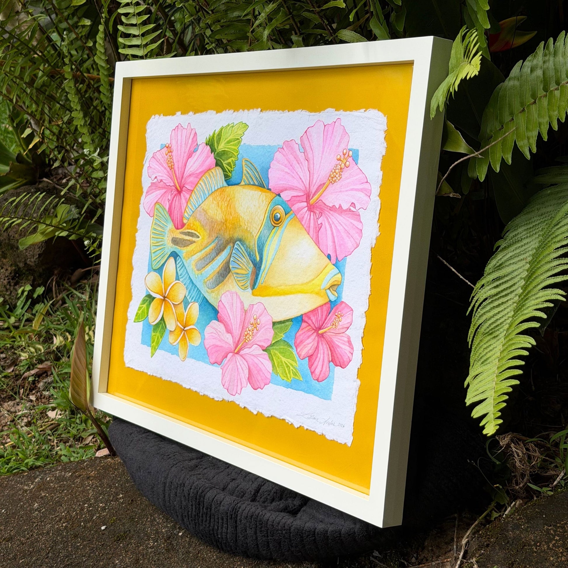 Framed artwork of a fish surrounded by pink flowers on a yellow background, leaning against green foliage. Framed watercolor painting of Humuhumunukunukuapua'a artwork color ful yellow trigger fish and hibiscus botanical flowers on a yellow background, white frame Humu_Harmony_2026_Liliane_Avalos_watercolor_painting_framed_Kauai_Hawaii_lifestyle
