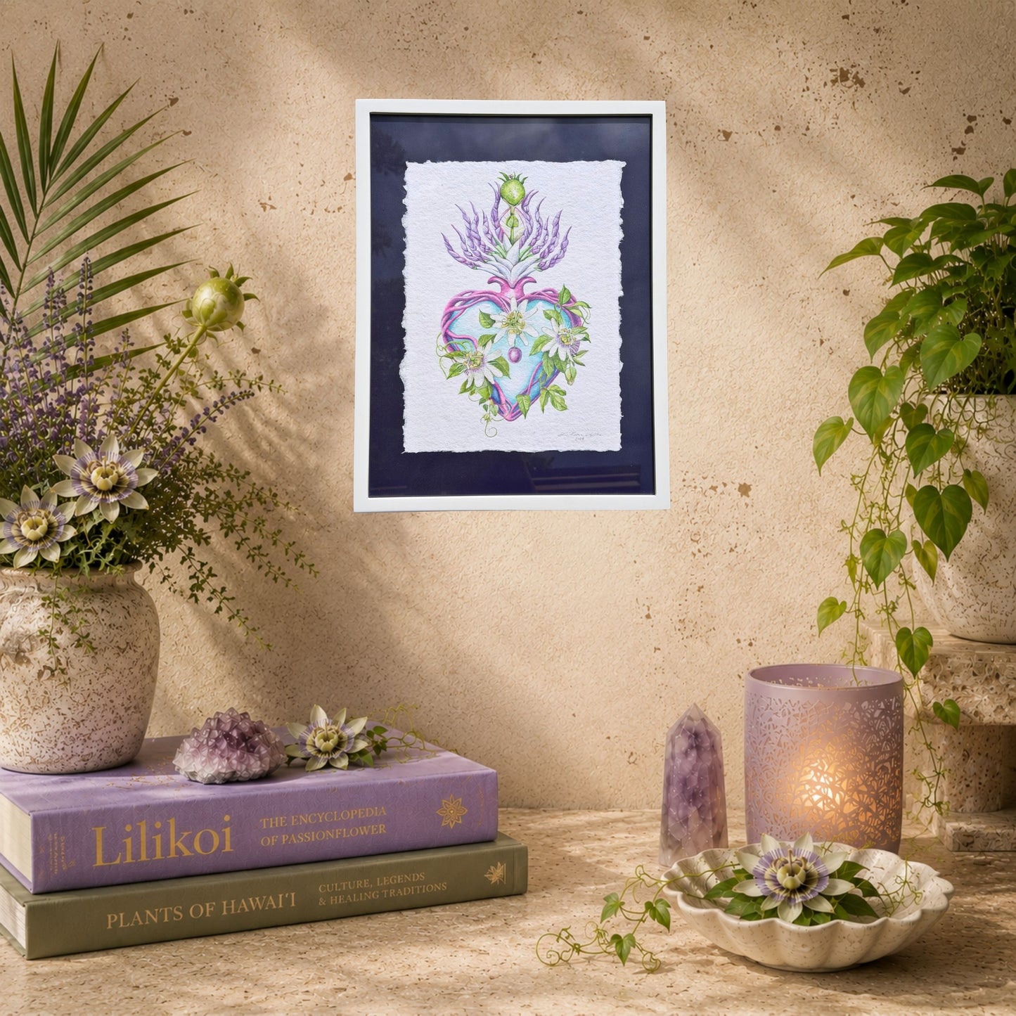 Framed artwork of a heart with flowers on a wall above books and decorative items. Framed original watercolor painting artwork with botanical sacred heart design featuring Lilikoi passion flowers and leaves on a white background of handmade paper, signed and embossed by artist Liliane Avalos