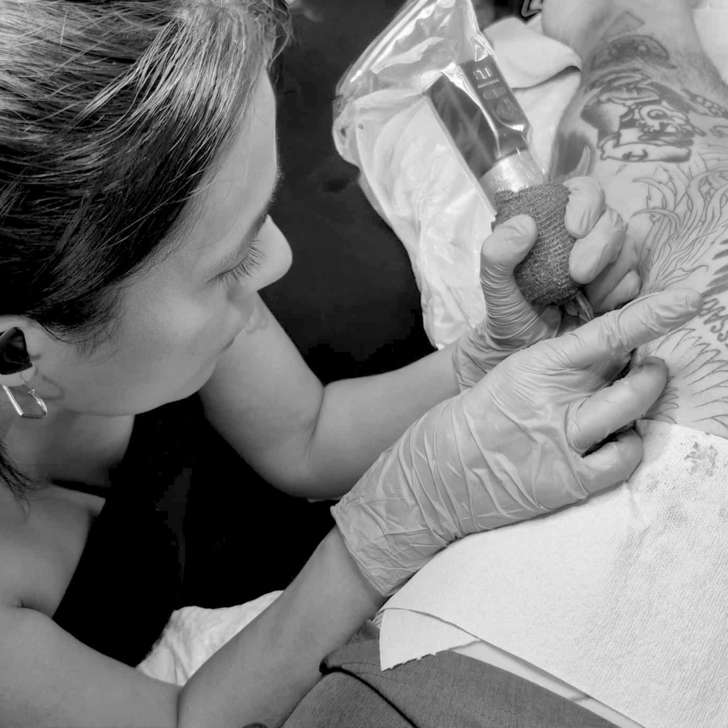 Kauai Hawaii tattoo artist Liliane Avalos with long dark hair and tattoos holding her hair, with illustrated hands in the background on a black background
Person getting a tattoo with a tattoo artist wearing gloves